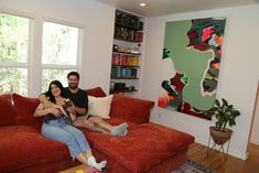 a man and woman sitting on a red couch in front of a window with bookshelves