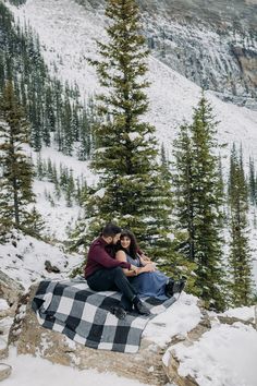 a man and woman sitting on top of a snow covered mountain next to evergreen trees