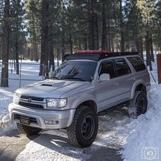 a silver truck parked on top of snow covered ground next to pine trees and fence