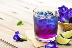 a glass filled with blue liquid next to purple flowers and lime slices on a wooden table