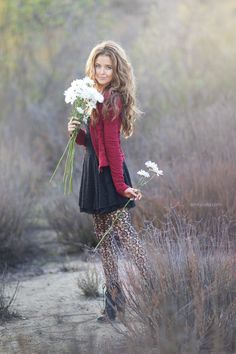 a woman holding flowers in her hands while standing on a dirt road with bushes and grass