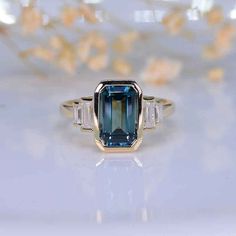 an emerald and diamond ring sitting on top of a white surface with flowers in the background