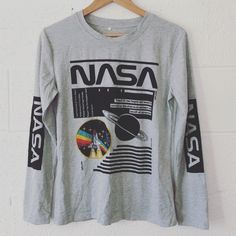 a gray nasa shirt hanging on a white wall with black writing and an image of saturn