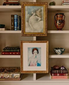 two framed pictures are sitting on shelves in front of books and vases, one with a woman's head