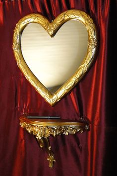 a gold heart shaped mirror sitting on top of a wooden shelf next to a red curtain