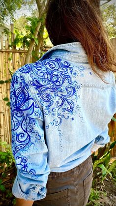 the back of a woman's shirt with an intricate blue design on it, in front of a bamboo fence