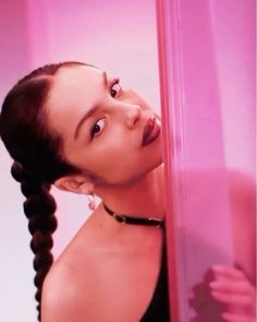 a woman with braids is leaning against a pink wall and looking at the camera