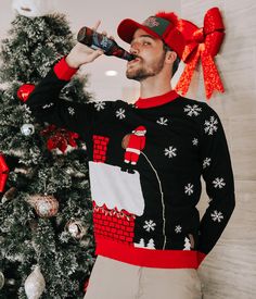 a man in an ugly christmas sweater drinking from a bottle next to a christmas tree