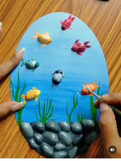 someone is painting a fish scene on a paper plate with rocks and water in the background