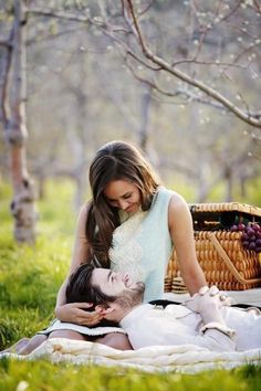 a man and woman laying on the grass in front of some trees, one holding his head