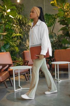 a woman in white shirt and pants walking through an indoor area with chairs, tables and potted plants