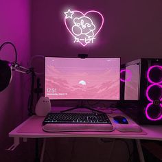 a desk with a keyboard, mouse and monitor in front of a neon heart on the wall