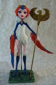 a doll with red hair and blue eyes holding a golden staff on a green base