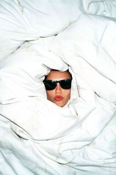 a woman wearing sunglasses hiding under a white comforter with her head covered by a blanket