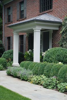 the house is surrounded by lush green bushes and shrubs, along with white pillars on either side