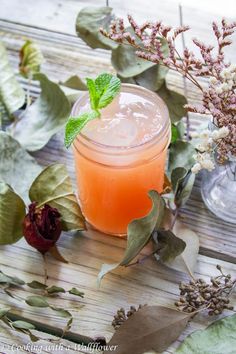 an orange drink sitting on top of a wooden table next to flowers and greenery