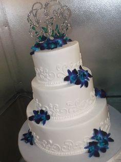 a three tiered white cake with blue flowers and a tiara on top is displayed