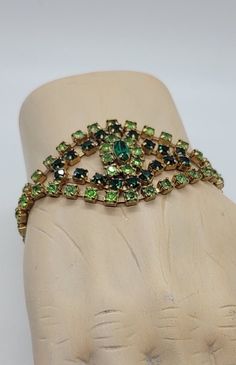 The beautiful rhinestone bracelet from the 1950s has a special open design and two colors of shiny green crystals. The vintage bracelet is made of 12K gold plated  metal, with a smooth, fluid feeling against the skin. But it is the design that makes it even more gorgeous. From the double row of prong-set lime green rhinestones mergers a center marquise-cut forest green stone. It is surrounded by light green crystals in a floral pattern and an outside frame of dark green round rhinestones.  On th Vintage Green Bracelets For Party, Vintage Green Jubilee Bracelet, Green Costume Jewelry Bracelets For Wedding, Green Rhinestone Bracelets For Wedding, Light Green Crystals, Vintage Bracelet, Rhinestone Bracelet, Green Crystals, Vintage Bracelets