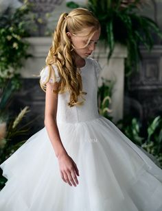 Crafted with impeccable attention to detail, the Teter Warm Couture Annie Girls Communion Dress is the epitome of elegance for a young lady's special day. This exquisite garment features a delicate lace cap-sleeve top that exudes a timeless charm, with a lace overlay bodice that adds a touch of sophistication. The back zip closure ensures a seamless fit, while the crystal-trim belt at the waist sparkles with every turn, highlighting the dress's exquisite craftsmanship. The back of the dress is a Baby Christening Outfit, Girls Communion Dresses, Girls Crown, Flower Girl Crown, Layered Tulle Skirt, Girls Couture, Christening Outfit, Girls Pageant Dresses, Crystal Trim