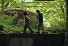 two boys walking across a bridge in the woods with an abandoned train car behind them