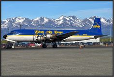 a blue and white plane is on the tarmac with mountains in the back ground