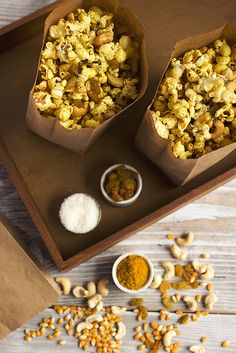 two brown paper bags filled with popcorn on top of a wooden tray next to bowls of nuts