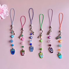 six different colored owl charms hanging from lanyards on a pink surface next to a flower