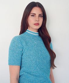 a woman with long brown hair wearing a blue shirt and jeans standing in front of a white wall