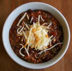 a white bowl filled with chili and cheese on top of a wooden table next to a spoon