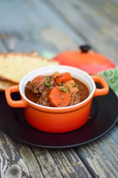 a bowl of stew on a black plate with bread and an orange pot next to it