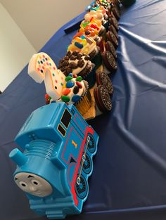 a toy train is lined up on a blue tablecloth with cookies and cupcakes