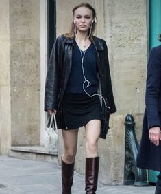 a woman walking down the street with headphones in her ears, wearing a black dress and knee high boots