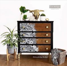 a black and white chest of drawers next to two plants on top of a wooden floor