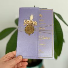 a person holding up a purple and gold wedding card with flowers on the front, in front of a potted plant