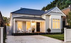 a white house with a black roof and two chairs in the front yard at dusk