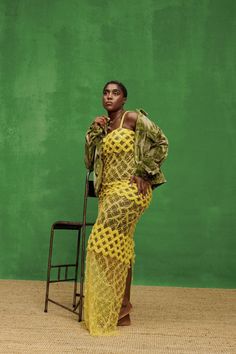 a woman standing in front of a green wall wearing a yellow dress and holding a chair