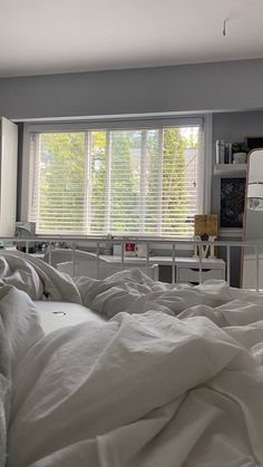 an unmade bed with white sheets and pillows in front of a window that has blinds