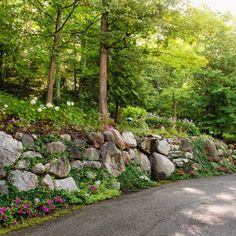 a stone wall with flowers growing on it next to a road in the middle of a forest