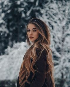 a woman with long hair standing in front of snow covered trees and looking off into the distance