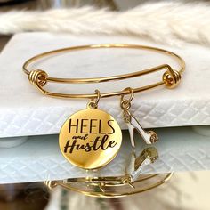 "Give A Girl The Right Jewelry And She Can Conquer The World\". Our mission is to provide products and gifts that will inspire, encourage and allow women to be their most confident selves. * Stainless Steel Gold Adjustable Bangle * Stainless Steel Gold Quote Charm \"Heels & Hustle\"  * Heel Charm * Hypoallergenic, lead and nickel free * Fits Average Size Wrist (approx. 7 inches) * Arrives beautifully packaged in our lovely white gift box. Includes a black cotton polishing cloth and cream jewelry pouch." Cream Jewelry, Quote Bracelet, Gold Quotes, Charming Quotes, Bracelet Quotes, Adjustable Bangle, Engraved Bracelet, Stackable Bracelets, Charm Bangle