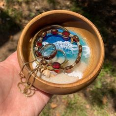 a hand holding a small wooden bowl with rings and beads in it on the beach