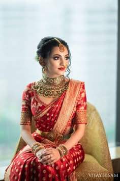 a woman in a red and gold sari sitting on a chair with her hands clasped to her chest