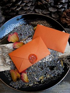 two orange envelopes sitting on top of a plate next to dried flowers and leaves