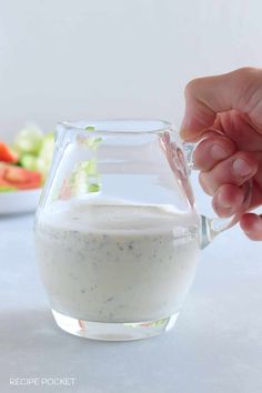 a person is pouring dressing into a glass pitcher on a table with vegetables in the background