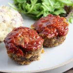 two meat patties covered in ketchup sitting on a plate next to mashed potatoes