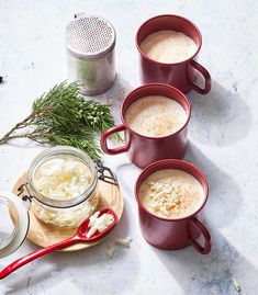 three red mugs filled with hot drinks next to spices and herbs on a table