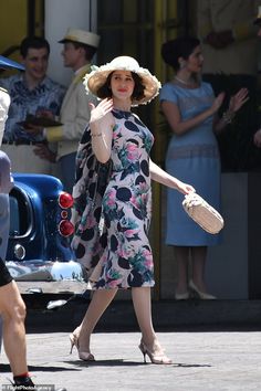 a woman in a floral dress and hat walking down the street with her hand on her hip