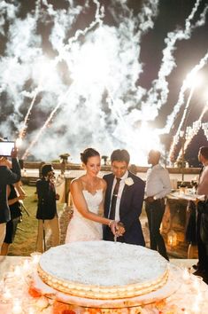 a newly married couple cutting their wedding cake with fireworks in the sky behind them and onlookers watching