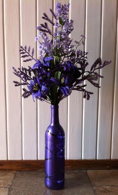 a purple vase filled with flowers on top of a stone floor next to a wall