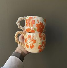 two coffee mugs with orange flowers on them being held by a person's hand